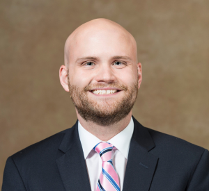 A bald man wearing a black suit jacket over a white collared shirt with a pink and blue striped tie.