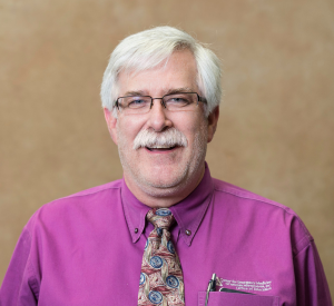 A man with white hair and a mustache wearing black-rimmed glasses, a pink collared shirt and a tie with pink, blue and tan patterns.