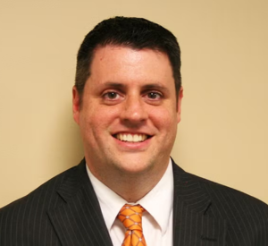 Man with short black hair wearing a black suit jacket over a white collared shirt and an orange tie.