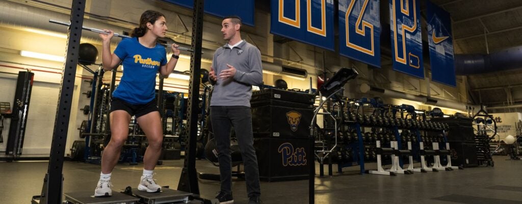 Female student athlete on a pressure plate under a weight bar talking to a sports scientist in a gym.