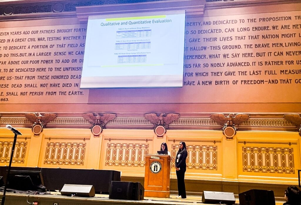 Two women stand at a podium on a stage with ornate decorations and a screen featuring their presentation