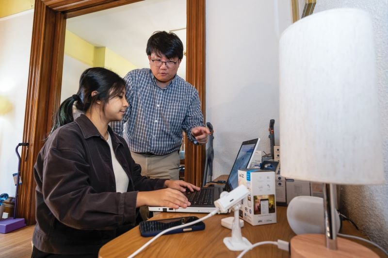 A man with dark hair wearing a gray collared shirt and khaki pants talking to a woman with dark hair pulled back wearing a brown sweater who is sitting at a desk.