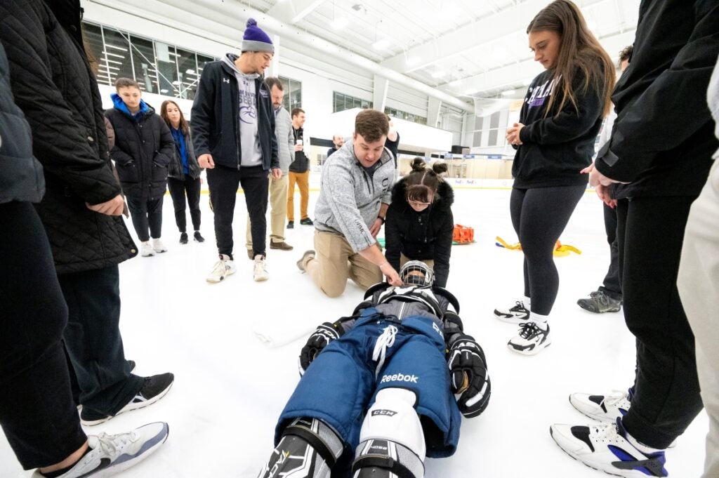 An instructor shows a student how to steady the head and neck of an ice hockey player laying on the ice