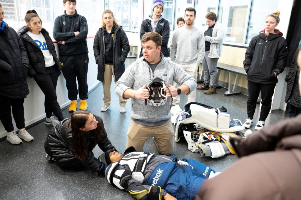 An instructor holds a hockey helmet as he demonstrates to students how to take care of a hockey player on the floor