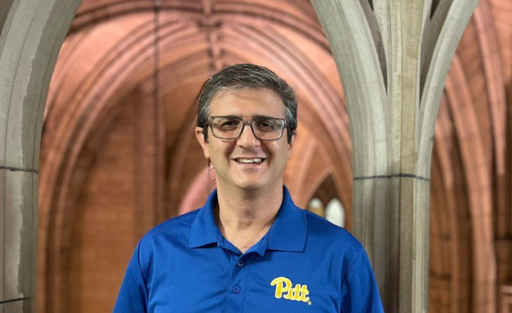 A man with short dark and grey hair wearing glasses and a blue polo shirt with the Pitt logo