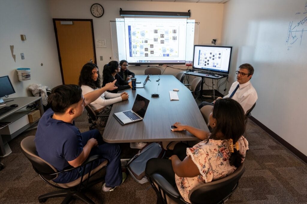 Five students and a male professor sit around a table in front of a presentation screen showing a data model