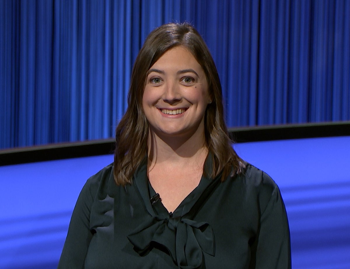 A woman with long dark hair wears a black blouse in front of a bright blue backdrop