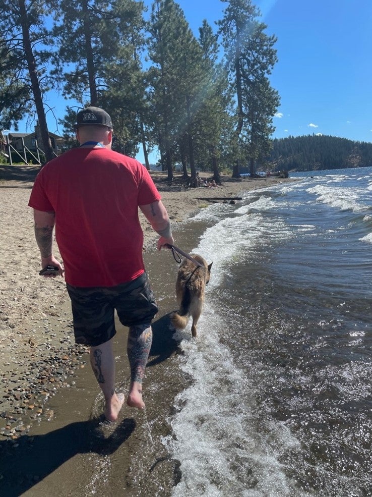 A man in a red shirt, shorts and hat walks a large tan dog along a lakeshore