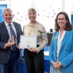 A man in a blue suit holds a glass award in his right hand, standing next to a woman with blond hair holding a certificate, and a third woman wearing a blue blazer