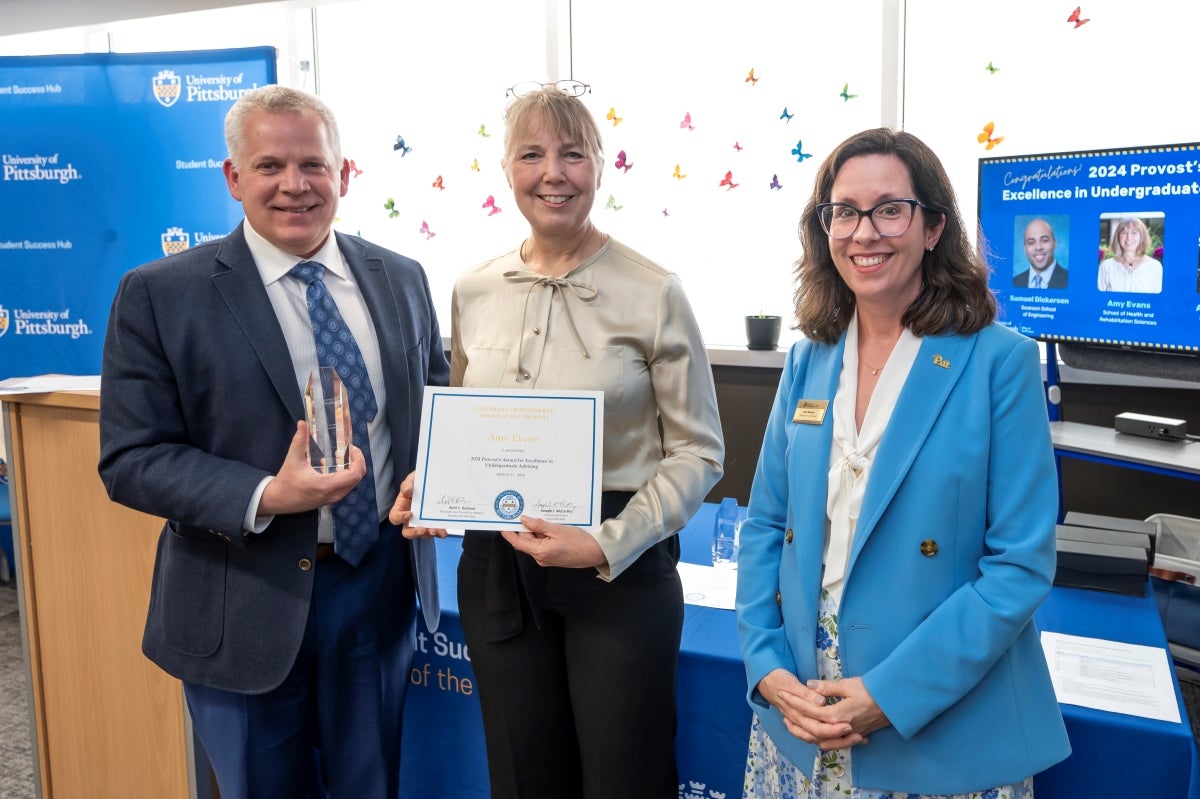 A man in a blue suit holds a glass award in his right hand, standing next to a woman with blond hair holding a certificate, and a third woman wearing a blue blazer