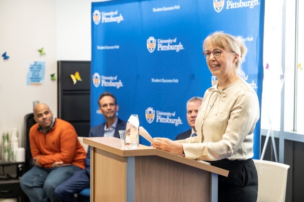 A woman with blond hair stands at a podium smiling with a glass award sitting on the podium, while three men in the background watch her speak