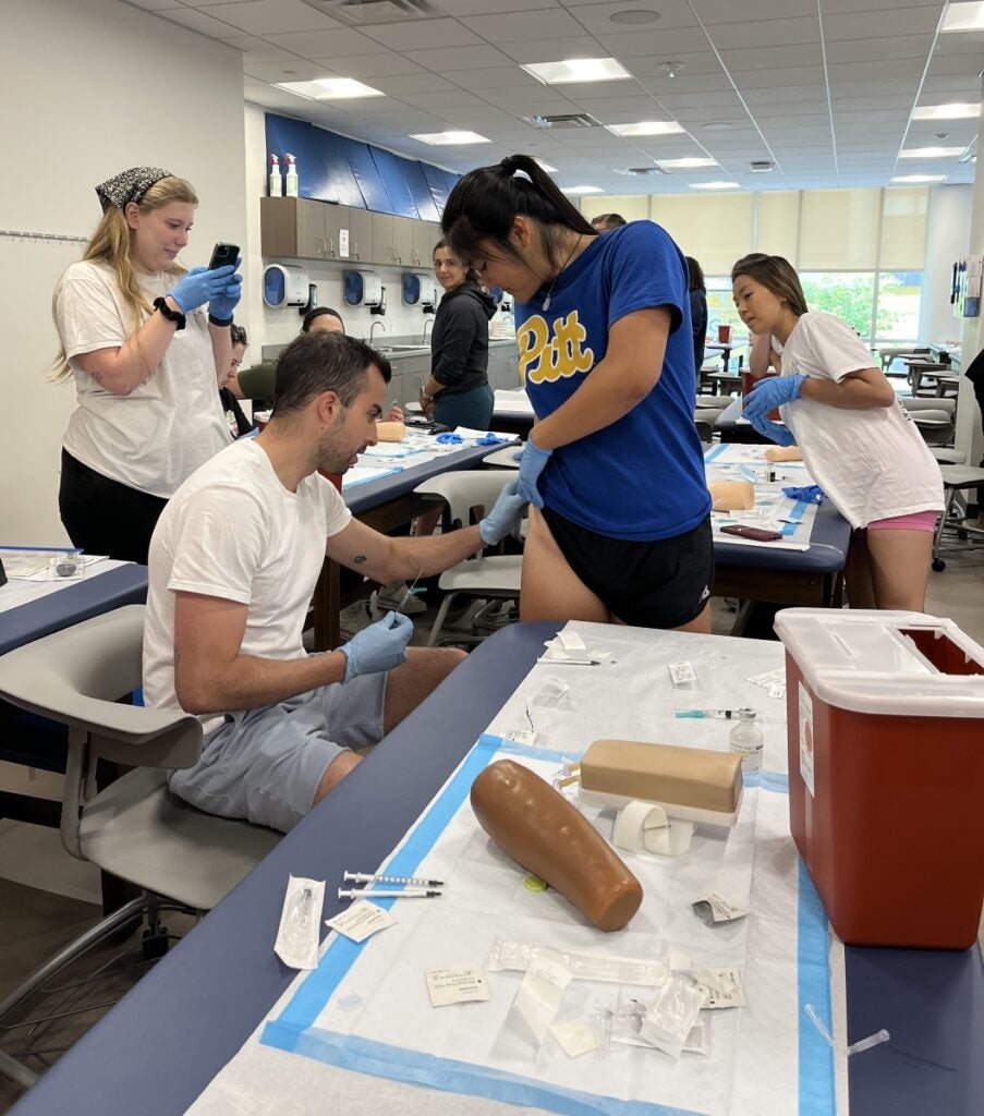 A woman in a blue Pitt shirt lifts the edge of her shorts so a male classmate sitting in a chair can clean an area for injection