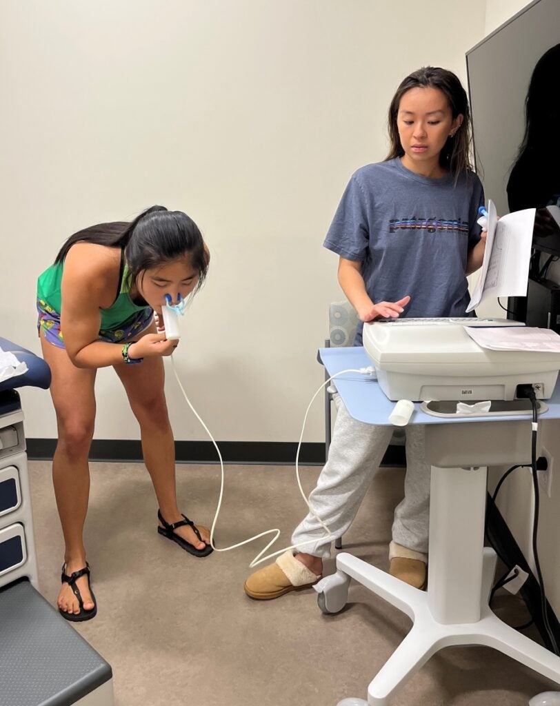 A woman leans over breathing into a tube connected to a machine monitored by another woman