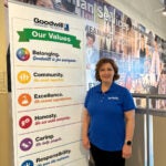 A woman with short auburn hair wearing a dark blue shirt and black pants standing next to a Goodwill sign.