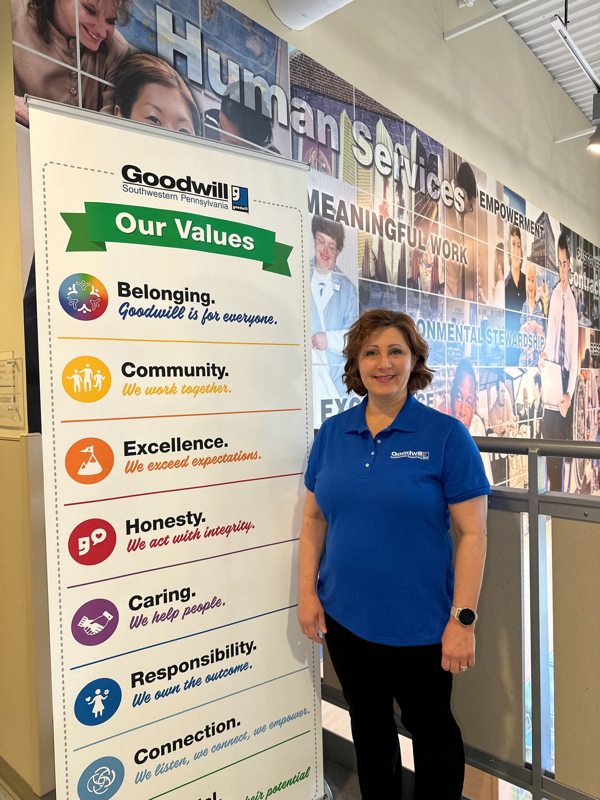 A woman with short auburn hair wearing a dark blue shirt and black pants standing next to a Goodwill sign.