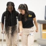 A woman practices walking with crutches while another woman on her left instructs her on the correct technique.