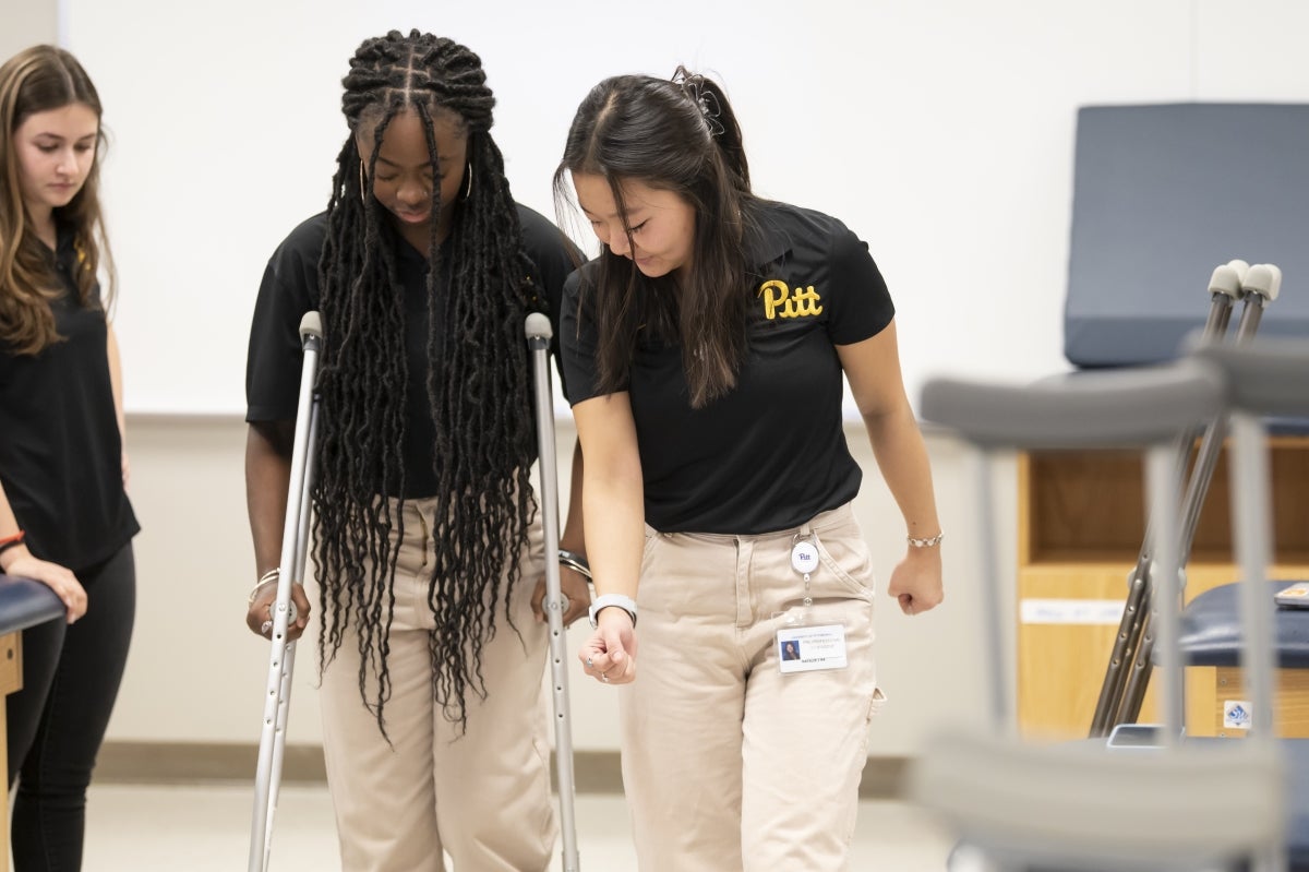 A woman practices walking with crutches while another woman on her left instructs her on the correct technique.