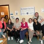 Six women sit in chairs and two women lean in behind them smiling. Behind them on the wall is a sign for the Birmingham Free Clinic