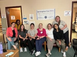 Six women sit in chairs and two women lean in behind them smiling. Behind them on the wall is a sign for the Birmingham Free Clinic