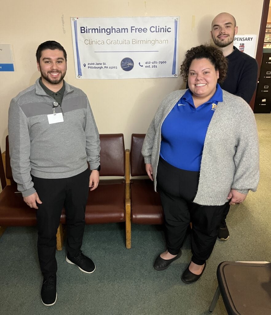 Three students stand smiling next to a sign for the Birmingham Free Clinic