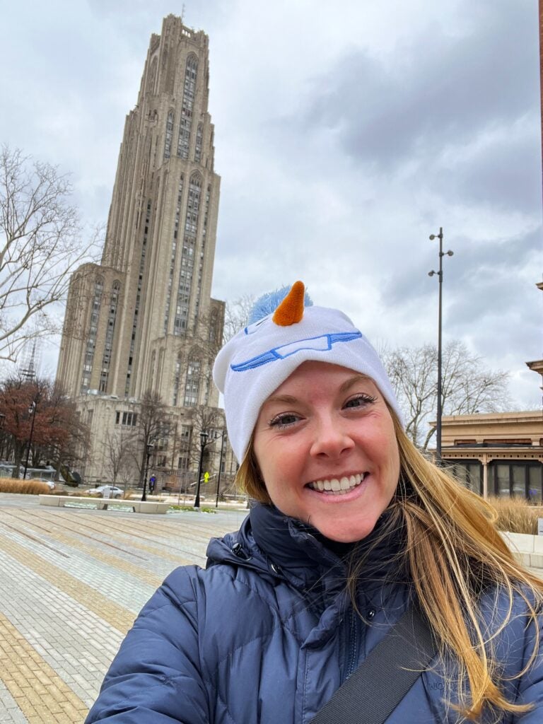 Woman with dark blonde hair wearing a white beanie and a black puffer jacket in front of the Cathedral of Learning.