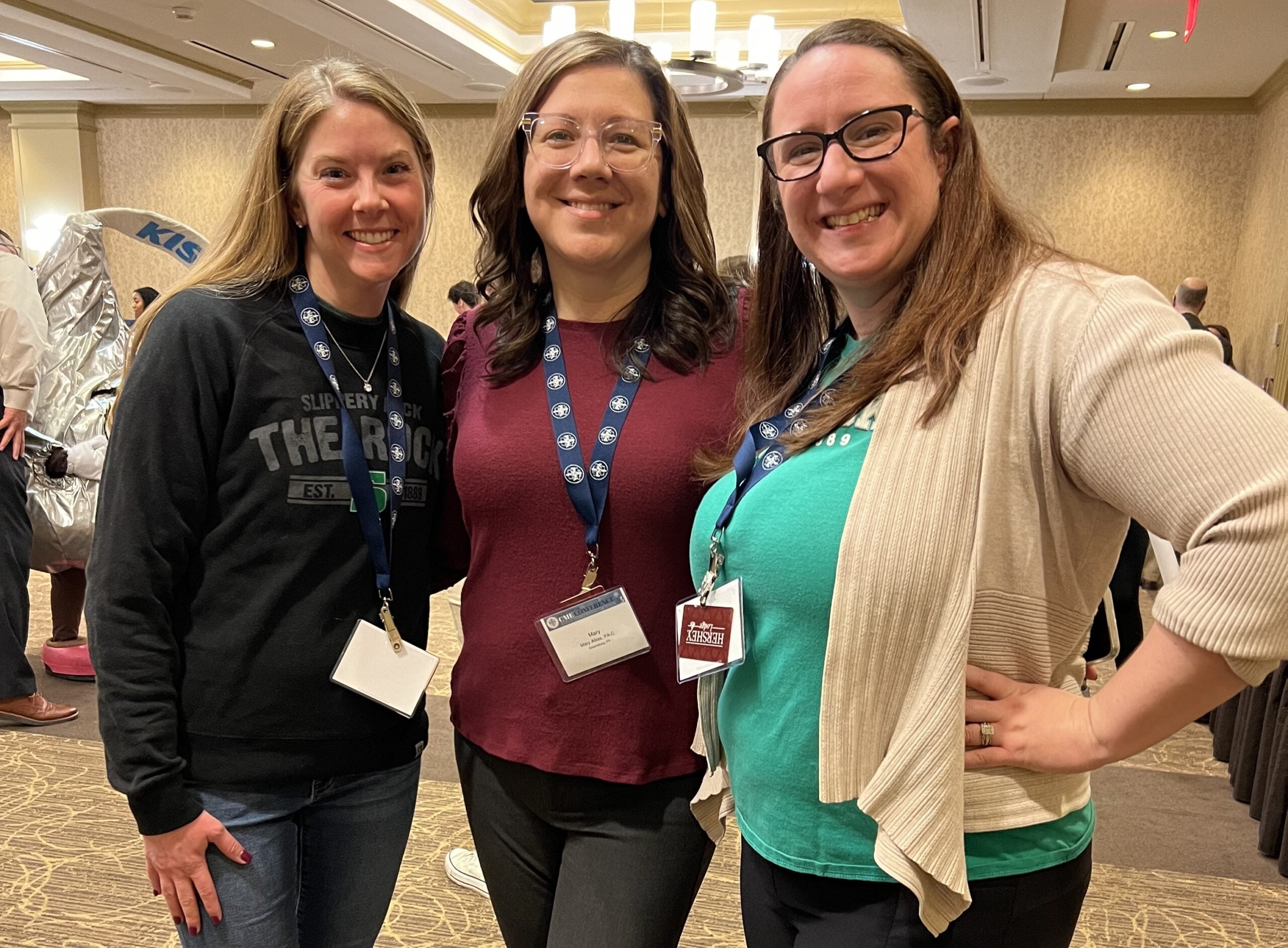 Three women standing together, all have brown hair. The woman on the left is wearing a gray sweater, the woman in the middle is wearing a red sweater and the woman on the right is wearing a white cardigan over a teal blouse.