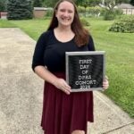 A woman with long dark hair in a black dress smiles and holds a sign that says, "First day of DPAS cohort 2024"