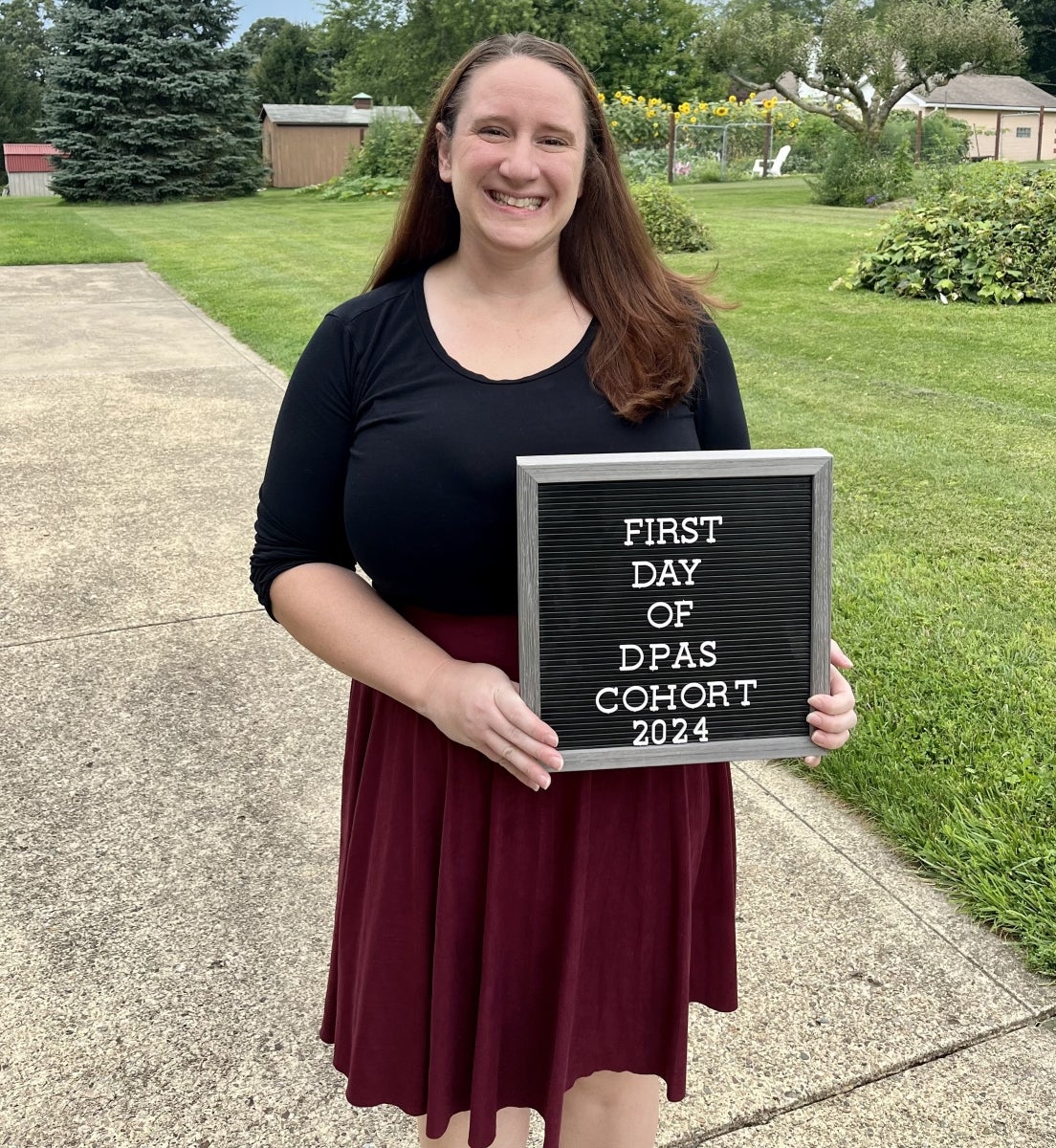 A woman with long dark hair in a black dress smiles and holds a sign that says, "First day of DPAS cohort 2024"