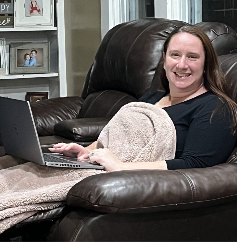 A woman with long dark hair reclines on a couch with a blanket and laptop computer