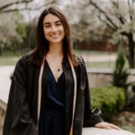 A woman with brown hair wearing a black graduation gown over a black dress.