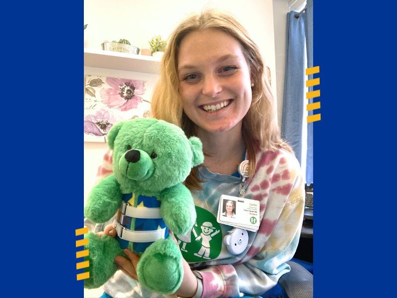 A woman with blonde hair holding a green teddy bear that wears a Thoracic-Lumbar-Sacral Orthosis around its torso.