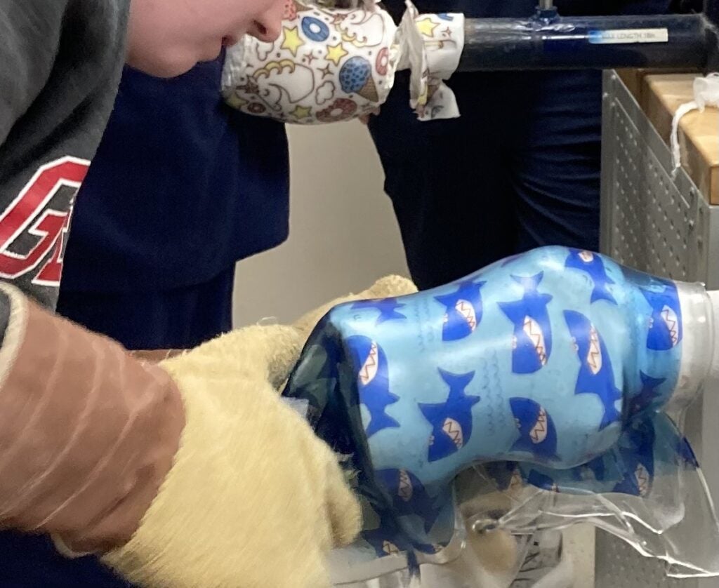 A student's hands are covered in heavy yellow gloves as he pulls clear hot plastic with a pattern of smiling cartoon sharks over a plaster mold.