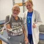 A woman with blonde hair wearing a white lab coat gives a thumbs up as she stands next to a man with white hair and a white beard and a prosthetic right arm.