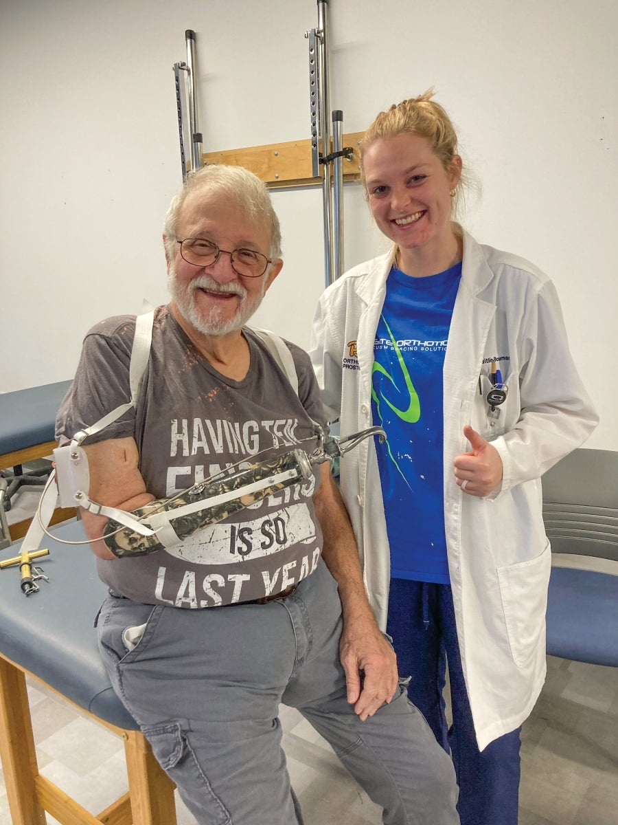 A woman with blonde hair wearing a white lab coat gives a thumbs up as she stands next to a man with white hair and a white beard and a prosthetic right arm.