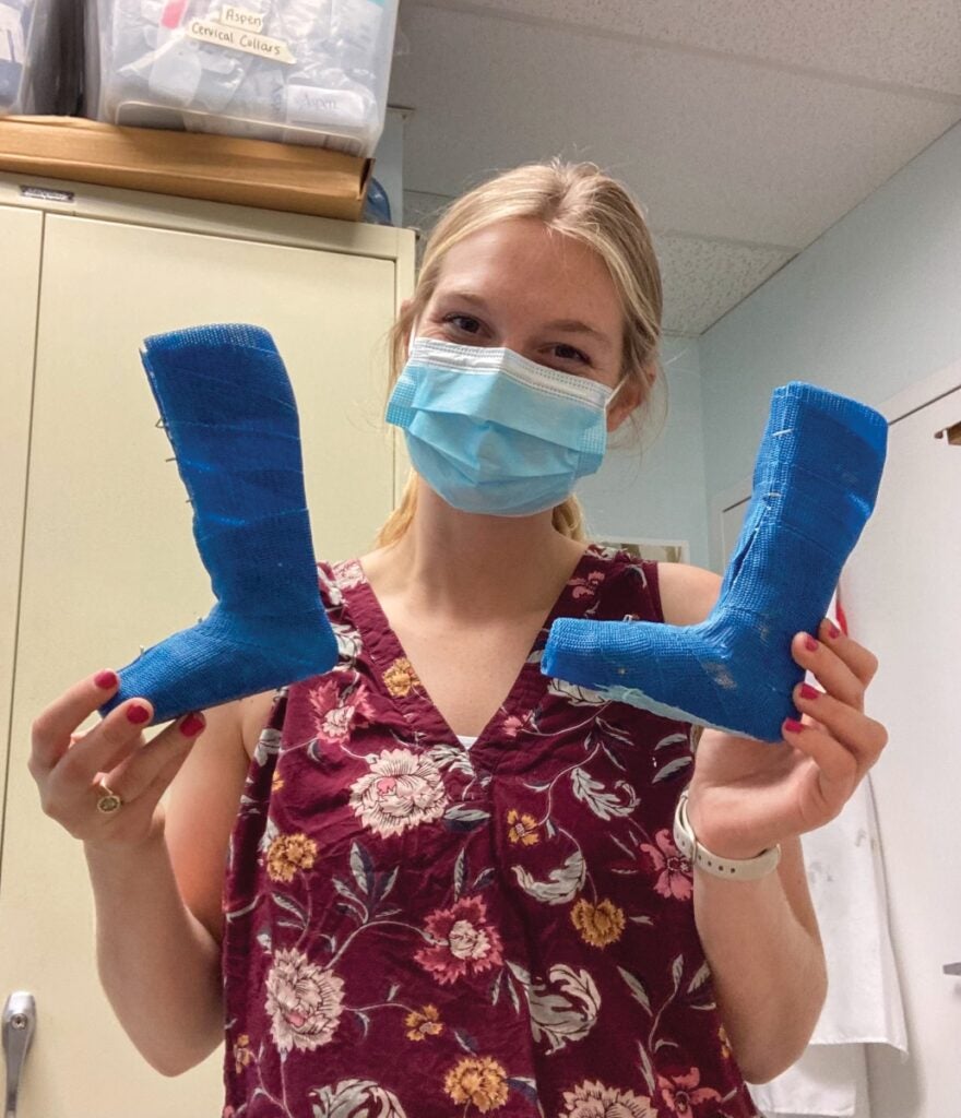 A woman with blonde hair and surgical mask holds up two small blue casts of a foot