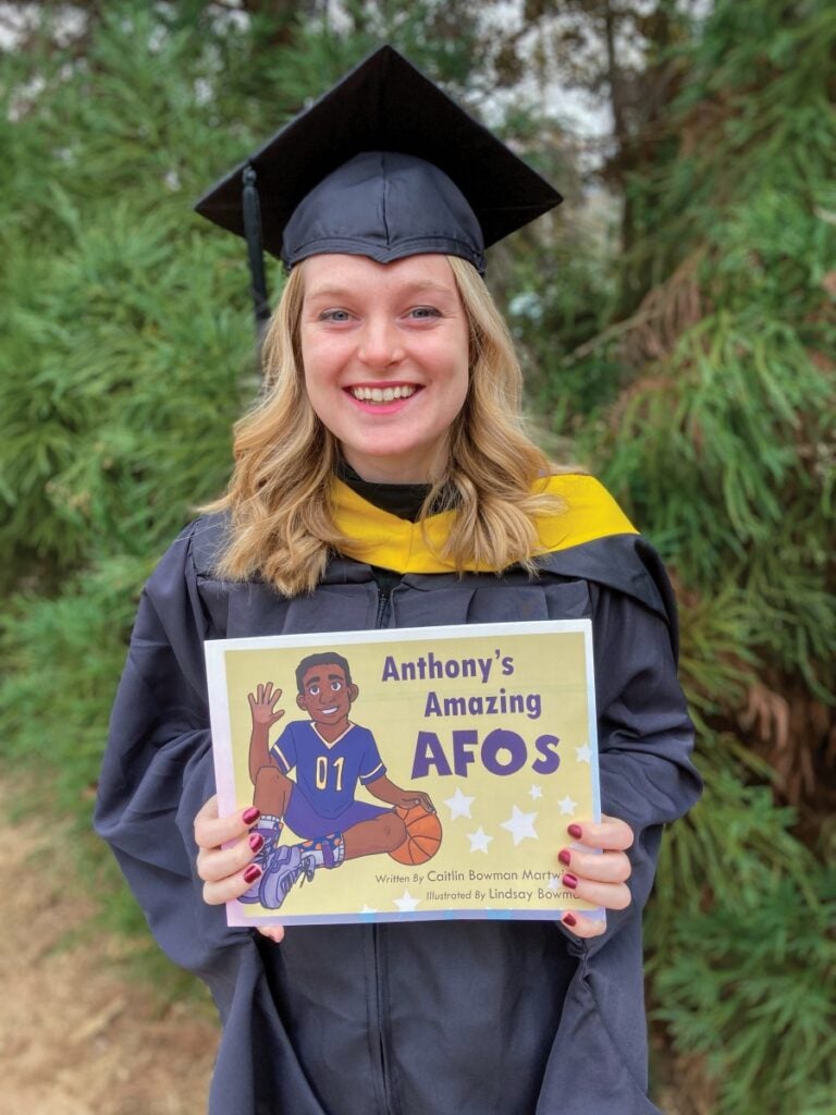 A woman with blonde hair wearing black graduation regalia smiles as she hold a book titled "Anthony's Amazing AFOs"