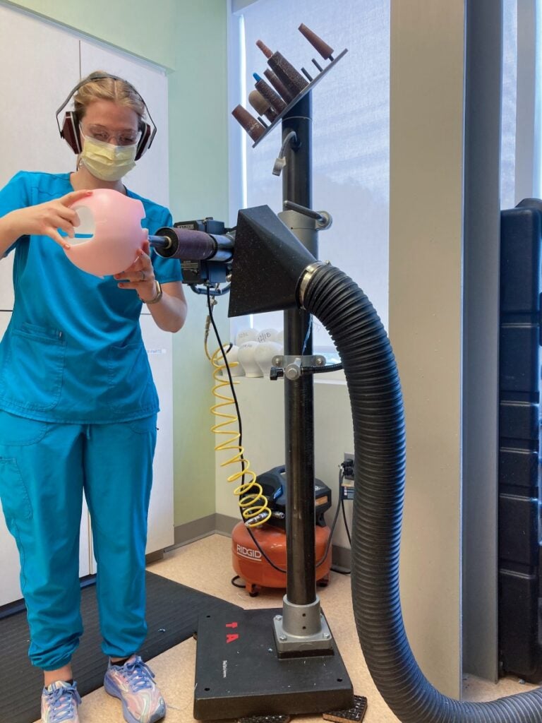 A woman in blue clothes wearing a mask holds a pink ball-shaped object against a large machine