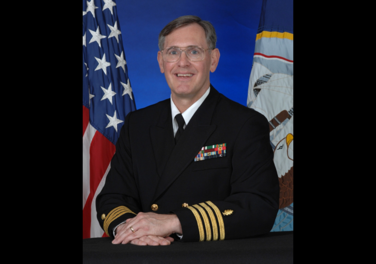 A man wears a formal black U.S. Navy coat with four gold bands on the sleeves and colorful military pins on the lapel.