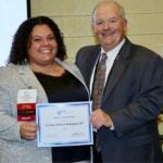 A woman with brown curly hair and a gray jacket smiles while holding a certificate with a man in a suit