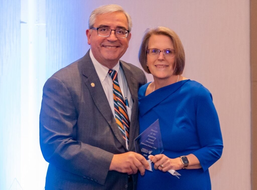 A woman with short blonde hair and glasses wearing a blue dress accepts a crystal award from a man with gray hair and glasses in a gray suit