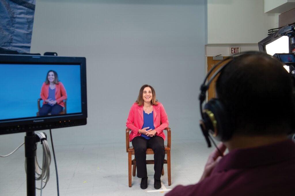 A woman wearing a bright pink jacket sits in a chair while being interviewed