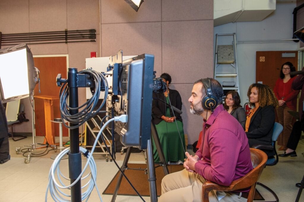 A man in a red short, dark short hair and a beard wears headphones and sits in a chair in front of video recording equipment