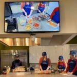 Four women stand around a kitchen work table cutting food and wearing blue Pitt Nutrition aprons and hats