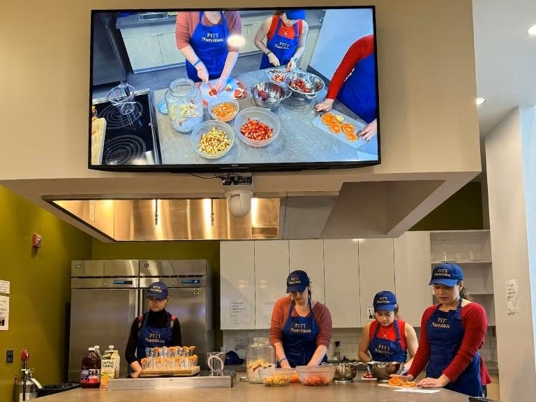 Four women stand around a kitchen work table cutting food and wearing blue Pitt Nutrition aprons and hats