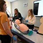 Students stand around a table with a plastic dummy to practice lab skills