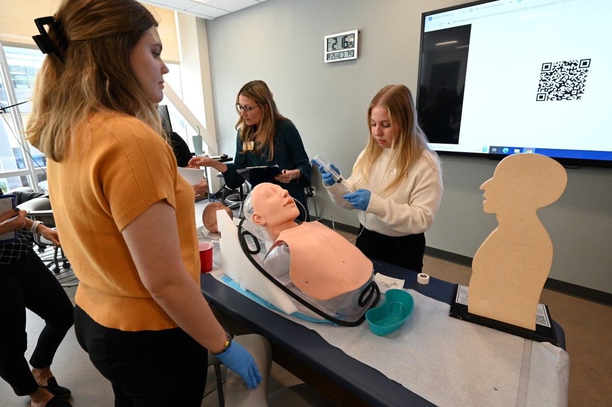 Students stand around a table with a plastic dummy to practice lab skills