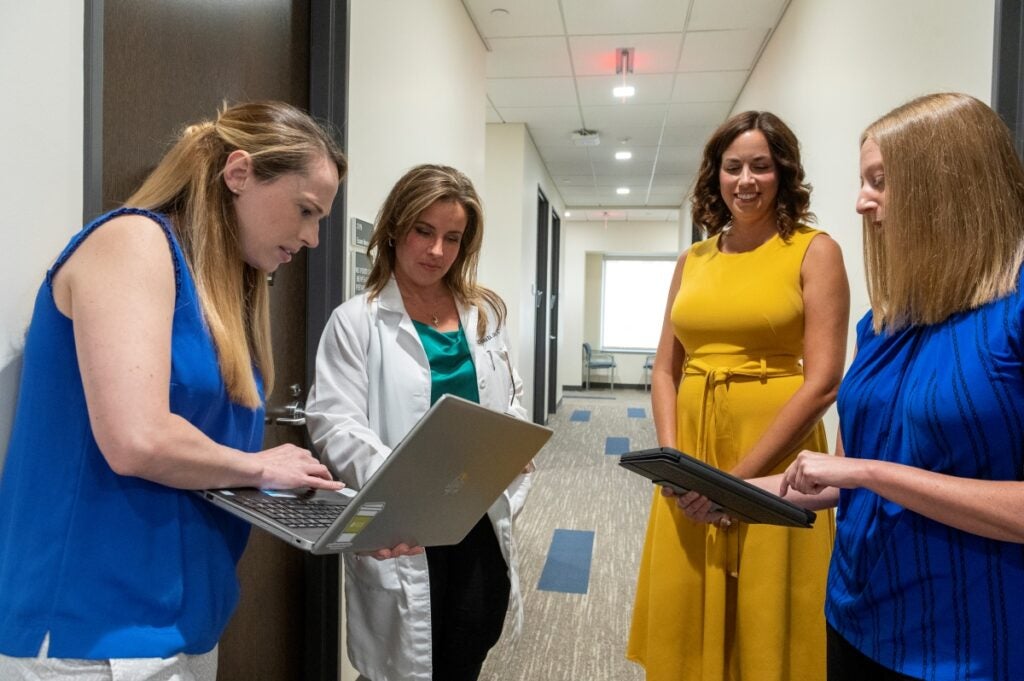 Four women stand in a hallway looking at information on laptop computers