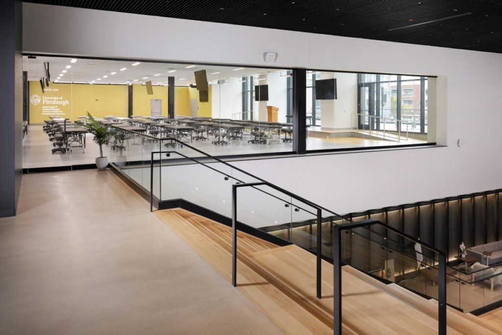 At the top of a wooden staircase, large windows open to a large classroom with patient tables and stools.