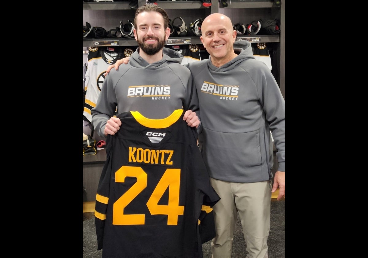 Two men wearing gray sweatshirts saying Bruins Hockey while the man with a beard hold a black hockey jersey with gold letters saying Koontz and the number 24