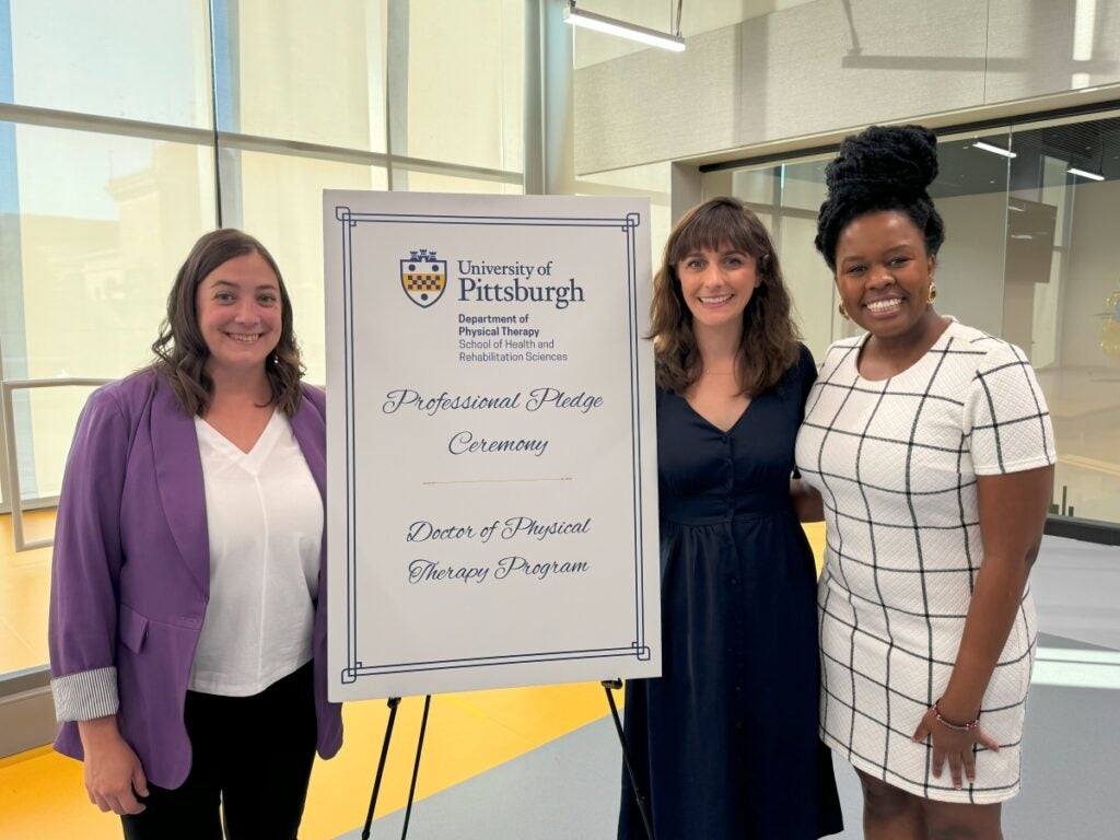 Three women stand and smile next to a sign for the Pitt Doctor of Physical Therapy Professional Pledge Ceremony